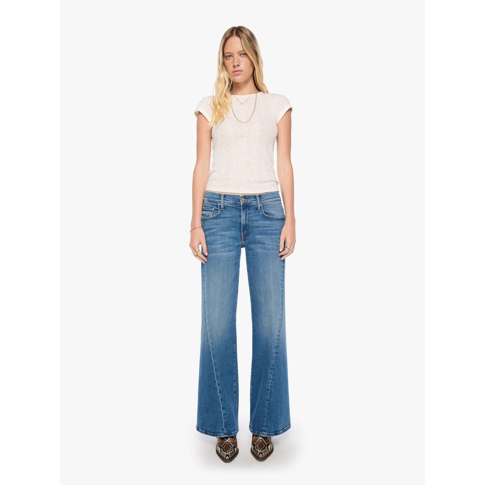 A woman modeling a pair of medium-wash blue wide-leg flare jeans with a textured cream short-sleeve top, standing against a plain white studio background.