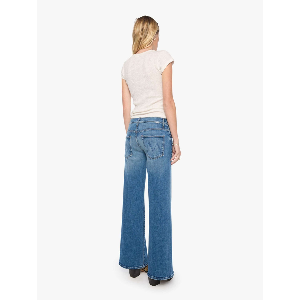 A woman viewed from behind wearing blue wide-leg jeans and a light cream short-sleeve top, standing against a plain light gray studio background.