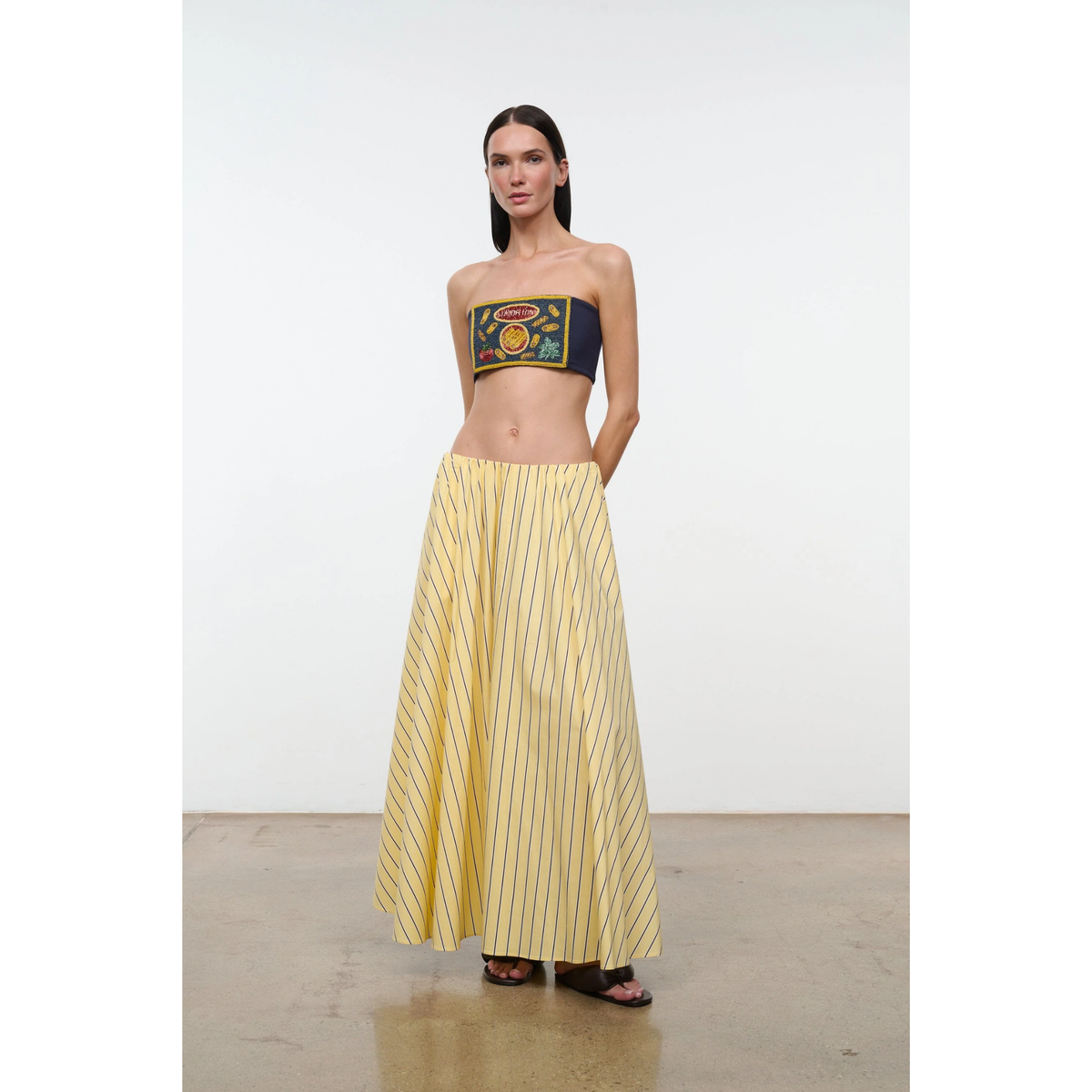 Woman wearing a strapless top with a colorful design and a long yellow skirt against a white background
