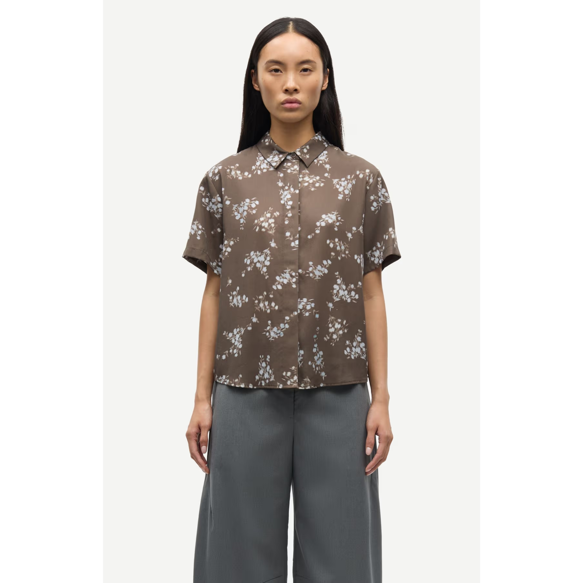A model wearing a brown short-sleeve button-up shirt with a white floral pattern, paired with gray wide-leg trousers, set against a plain light gray background.