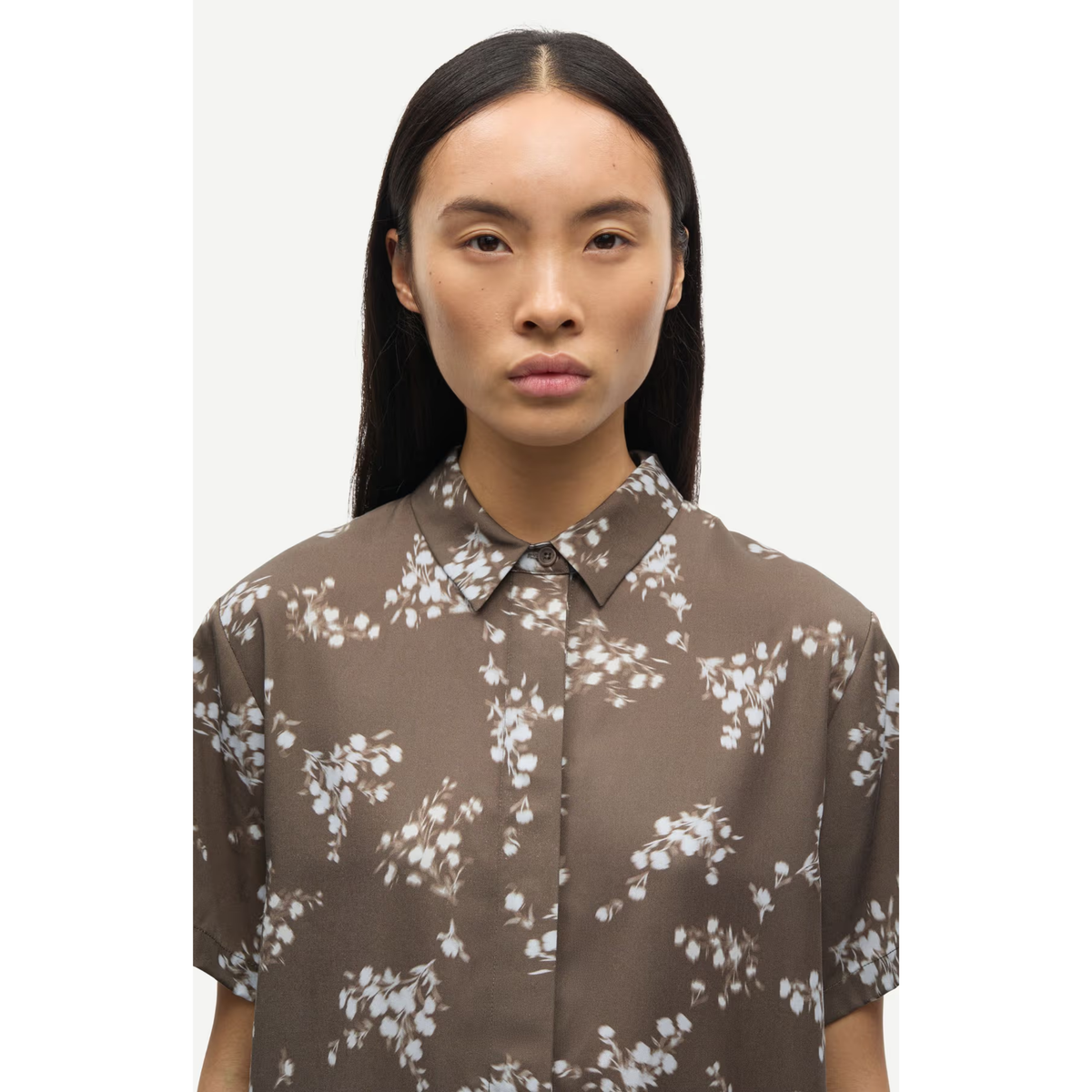 A woman with long, straight black hair wearing a brown short-sleeve button-up shirt with a white floral print, photographed against a plain light gray background.