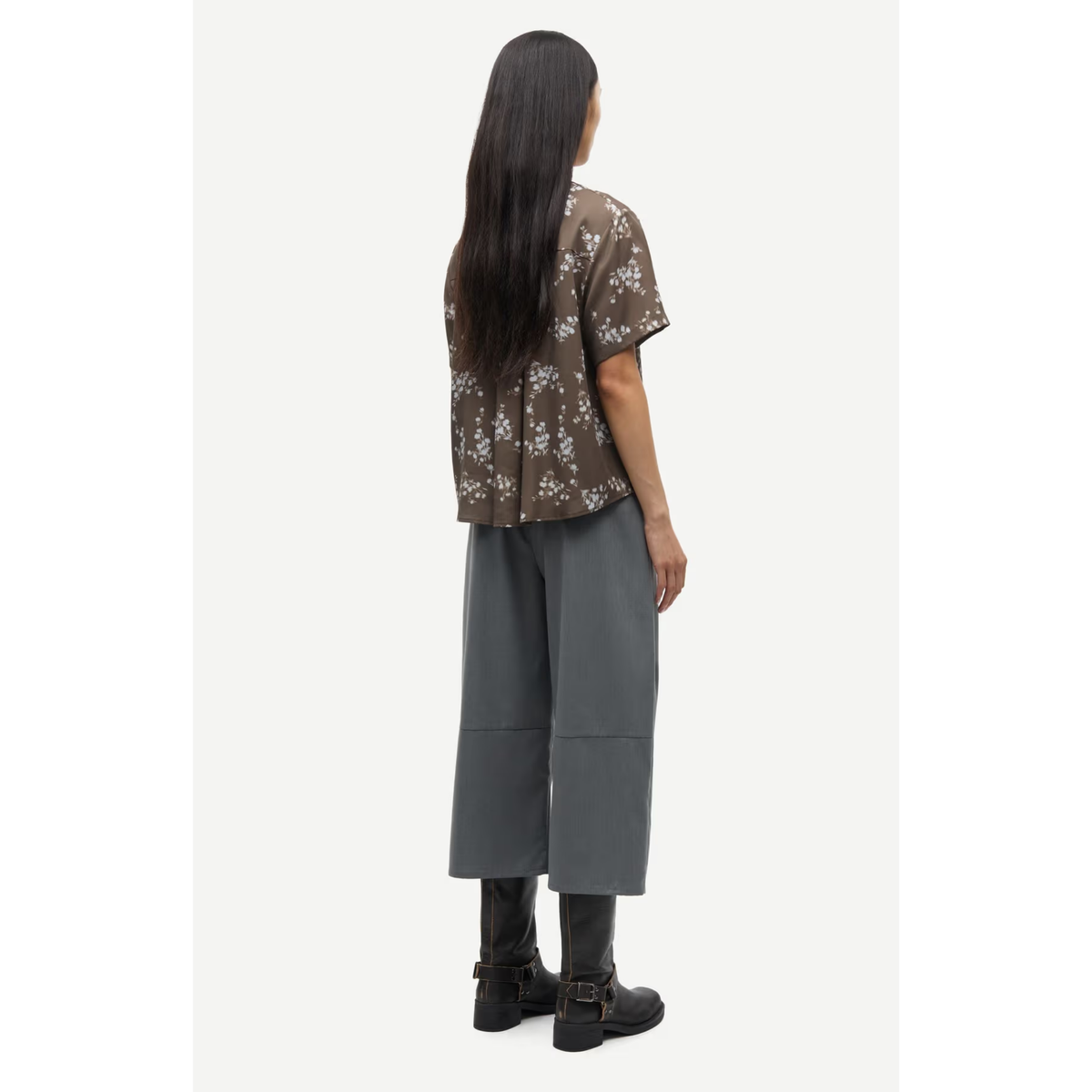A model shown from the back wearing a brown short-sleeve button-up shirt with a white floral pattern, paired with dark gray cropped pants and black boots, against a light gray studio background.