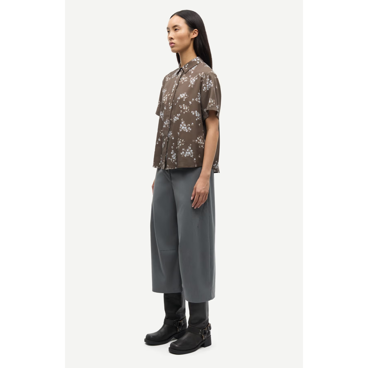 A model is shown in profile wearing a brown short-sleeve button-up shirt with a white floral pattern, paired with wide-leg gray trousers and black boots, against a plain light gray background.