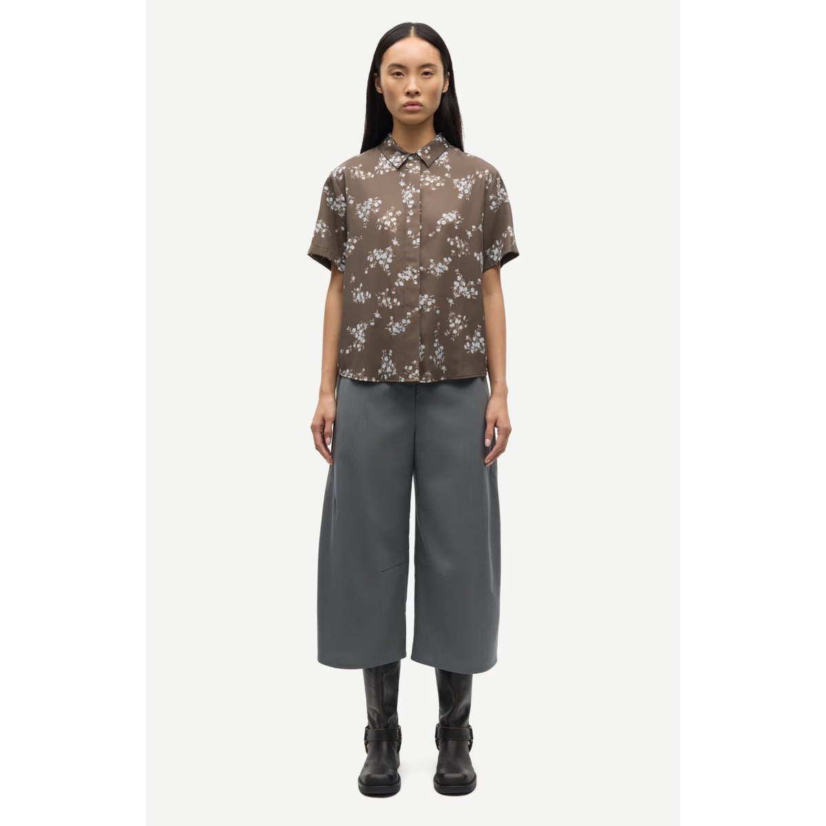 A model stands against a light gray background wearing a brown short-sleeve button-up shirt with a white floral pattern, paired with wide-leg gray trousers and black boots.