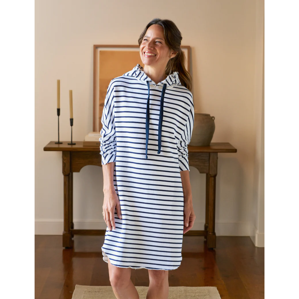 Woman wearing a white and blue striped dress with a hood in a room with wooden furniture and a painting.