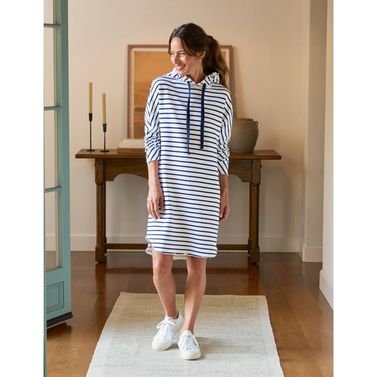 Woman wearing a striped dress standing in a room with wooden flooring and a table in the background.