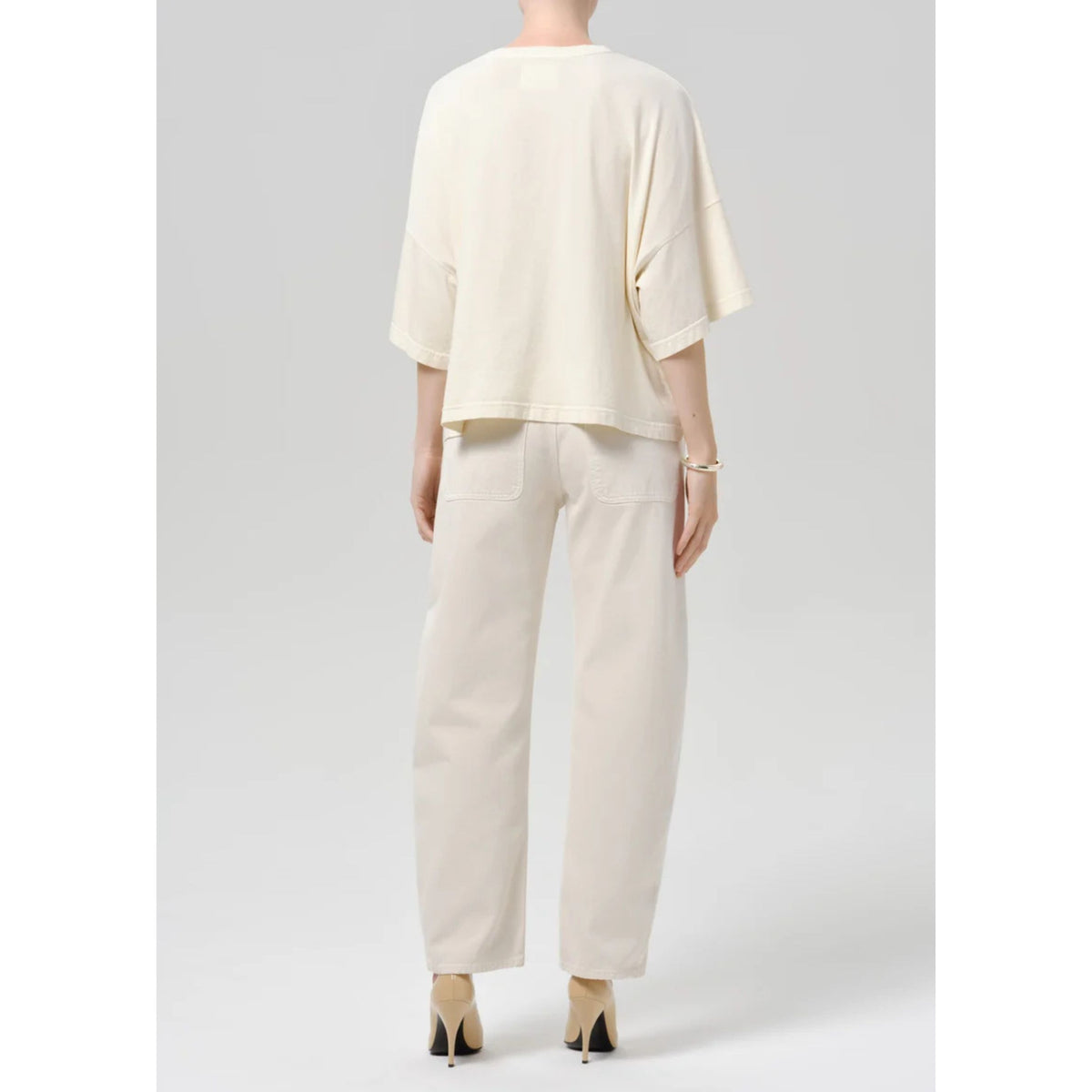 A model shown from the back wearing a cream-colored oversized short-sleeve top and matching cream wide-leg trousers, paired with beige high heels, against a light gray studio background.