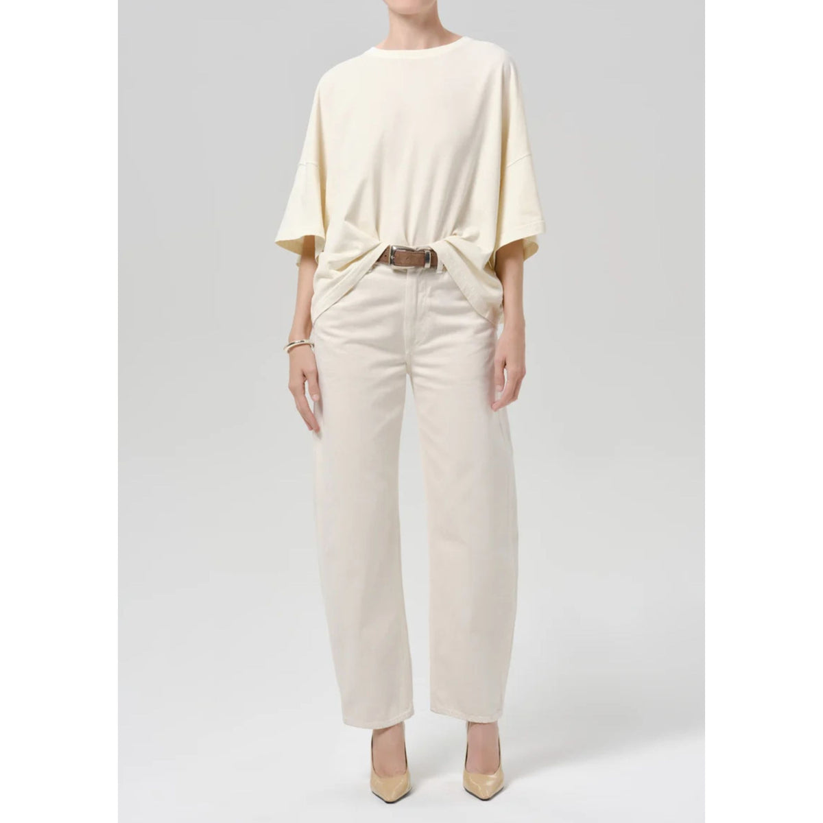 A model wearing a cream-colored short-sleeve top and matching wide-leg trousers, styled with a brown belt and beige heels against a light gray studio background.