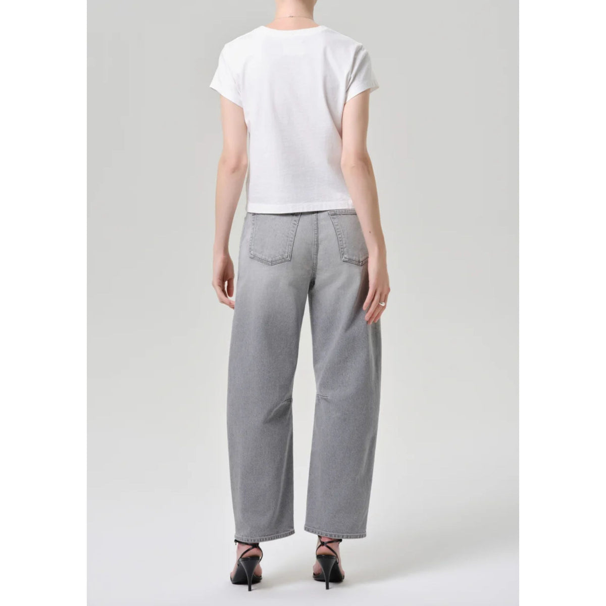 A model shown from the back wearing a white short-sleeve t-shirt and light gray wide-leg jeans, paired with black high-heeled sandals, against a light gray studio background.