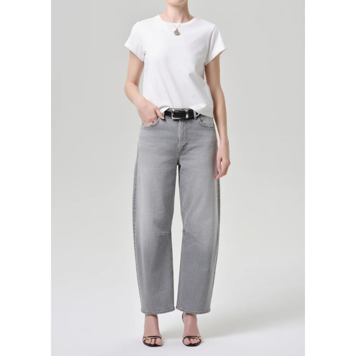 A model wearing a white short-sleeve t-shirt, light gray wide-leg cropped jeans with dark side stripes, a black belt, and black sandals, standing against a light gray studio background.