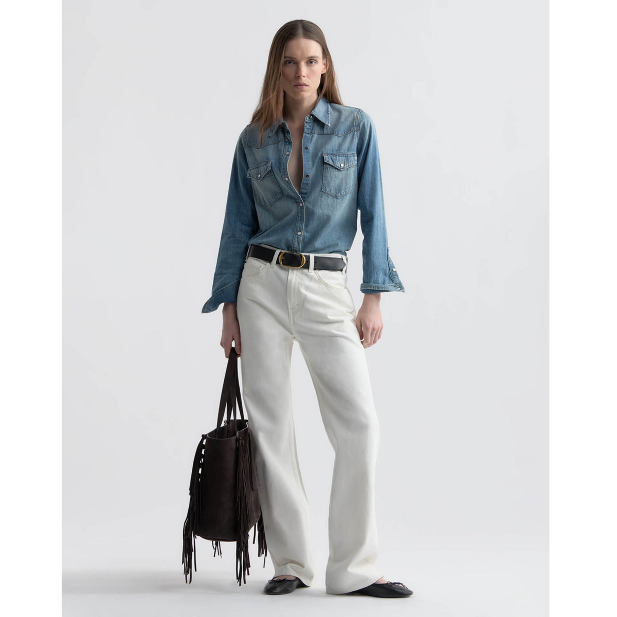 Woman wearing a denim shirt and white pants with a black handbag on a light gray background