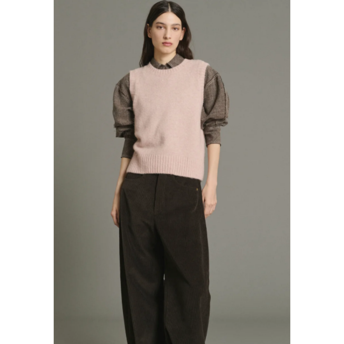 A model wearing a light pink ribbed knit sweater vest over sheer dark brown long-sleeve bubble sleeves, paired with dark brown wide-leg trousers, against a solid gray studio background.