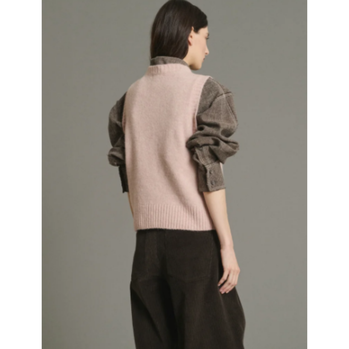 A model shown from the back wearing a light pink ribbed knit vest over a brown textured long-sleeve top and dark brown wide-leg trousers, set against a solid gray studio background.