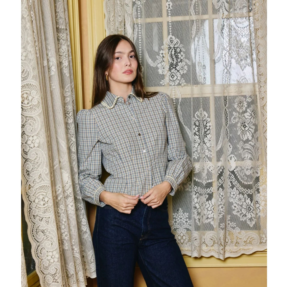 A woman stands indoors wearing a long-sleeve button-front blouse with a fine blue, beige, and white gingham check pattern, paired with dark blue jeans. She is posed in front of lace curtains and a window.