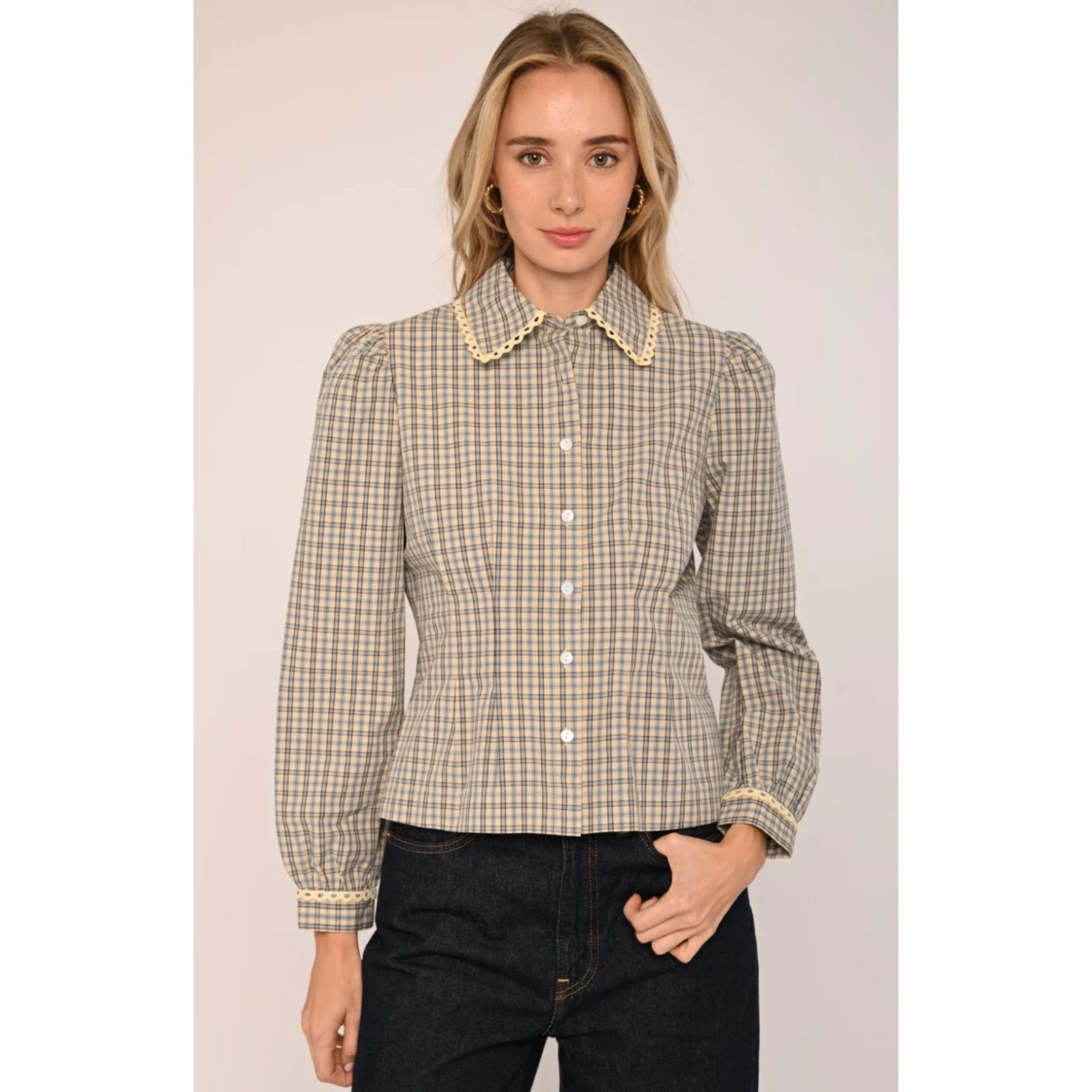 A woman modeling a beige and brown checkered long-sleeve blouse with a decorative pearl collar, paired with dark jeans, against a solid light beige background.