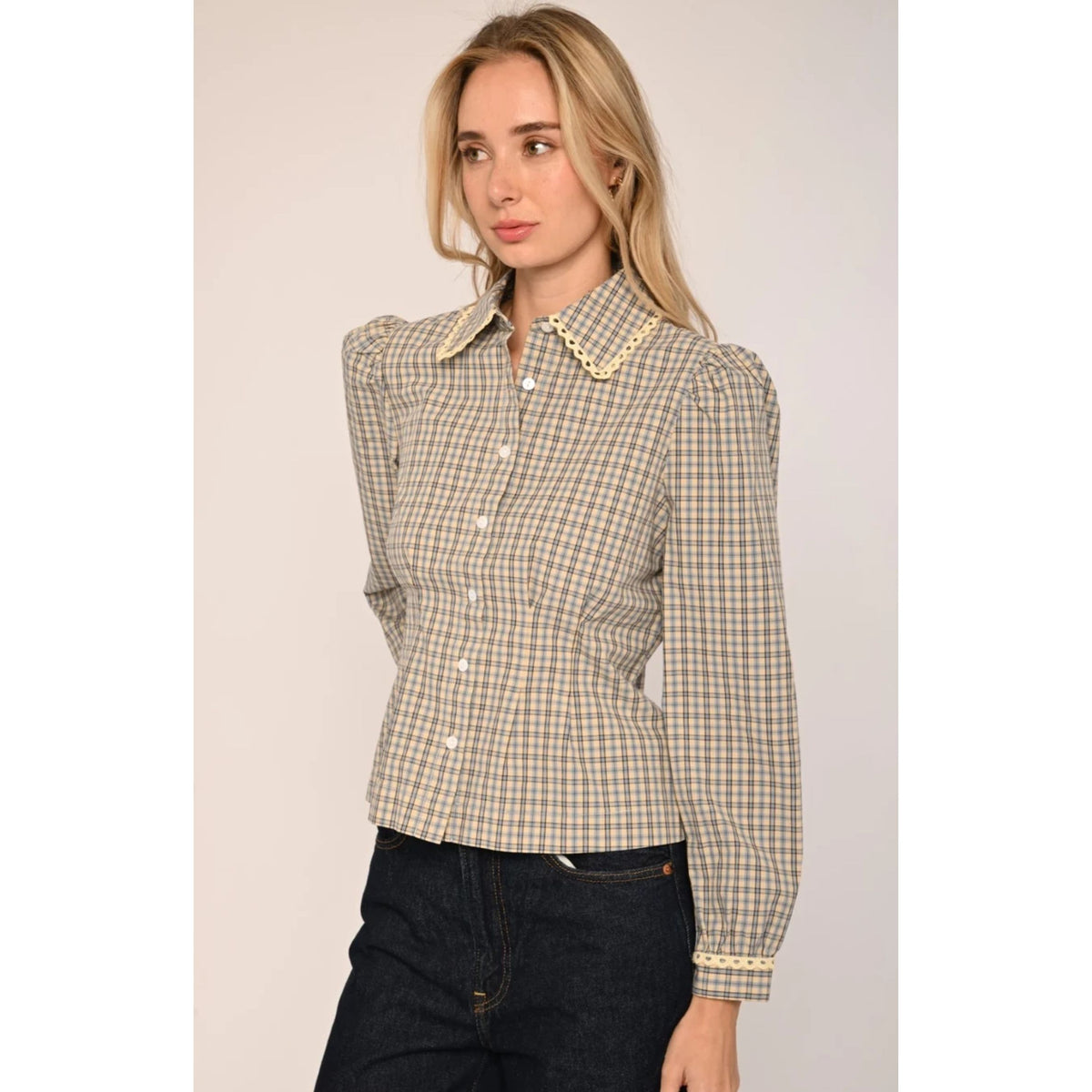 A woman modeling a beige and white checkered long-sleeve blouse with puffed shoulders and a decorative pearl collar, paired with dark jeans against a plain light background.