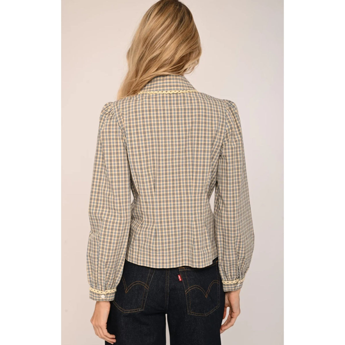 A woman shown from the back wearing a beige and brown plaid long-sleeve button-up shirt with decorative gold trim on the collar and cuffs, paired with dark jeans.
