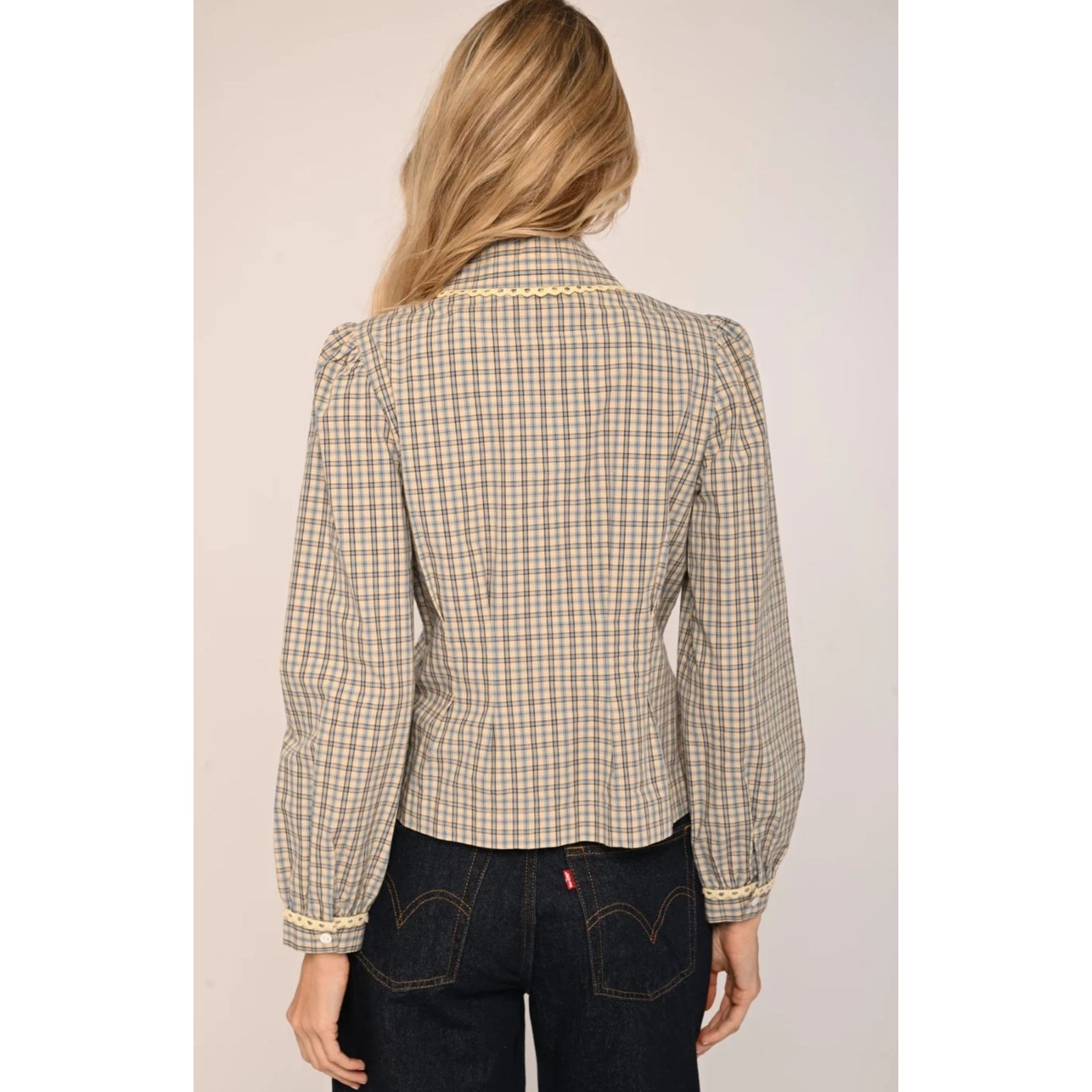 A woman modeling a beige and brown checkered long-sleeve blouse with a decorative pearl collar, paired with dark jeans, against a solid light beige background.