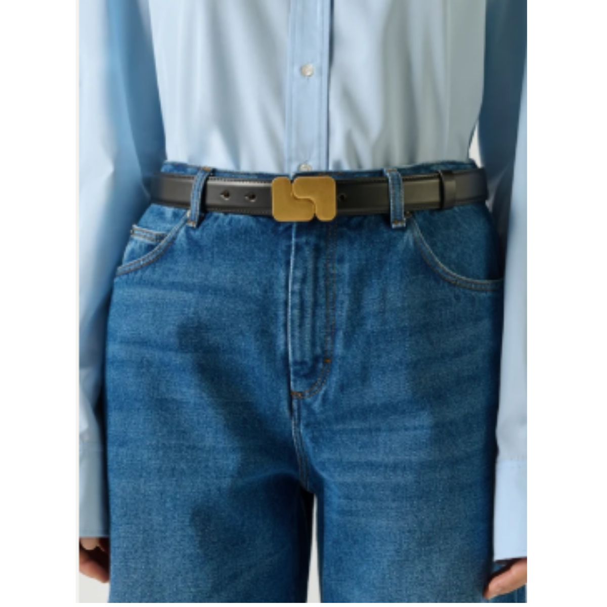 A close-up of a person wearing a dark brown leather belt with a gold-tone geometric buckle, styled with blue jeans and a light blue button-down shirt.