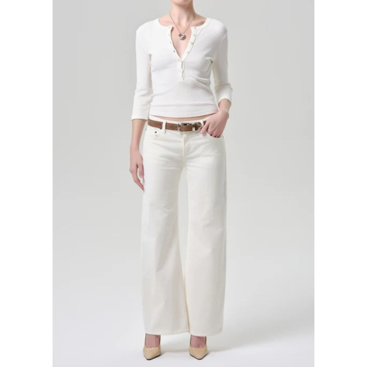A woman modeling a white long-sleeve henley top with a split neckline and white wide-leg pants, styled with a brown belt and beige heels against a light gray studio background.