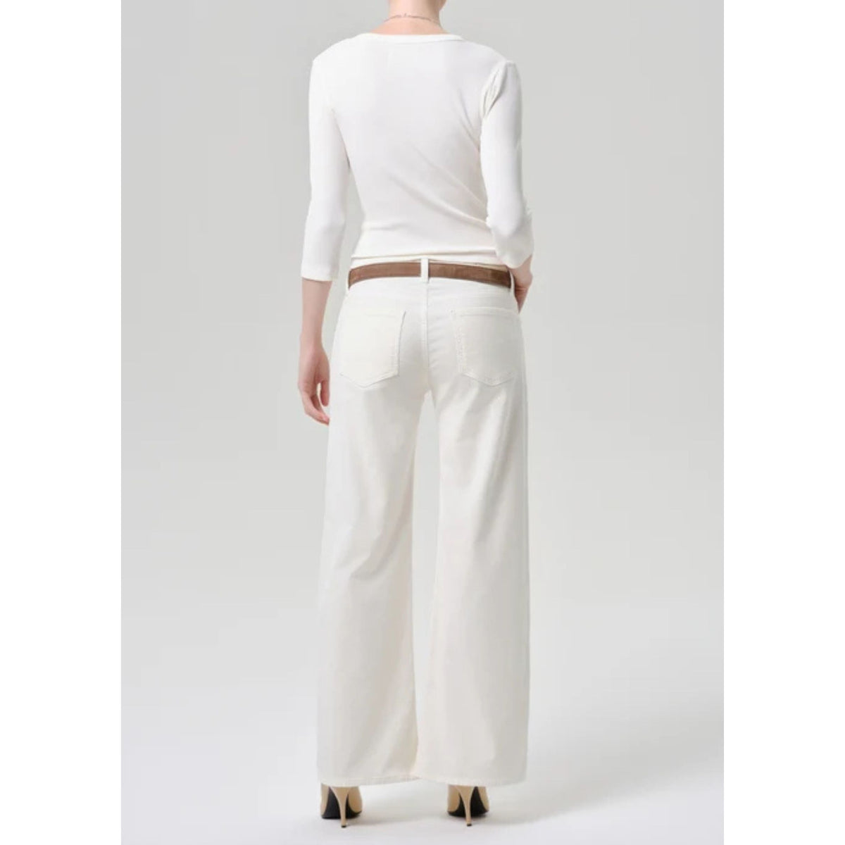 A model shown from the back wearing a white long-sleeve top, white wide-leg pants, and a brown belt, set against a plain light gray studio background.