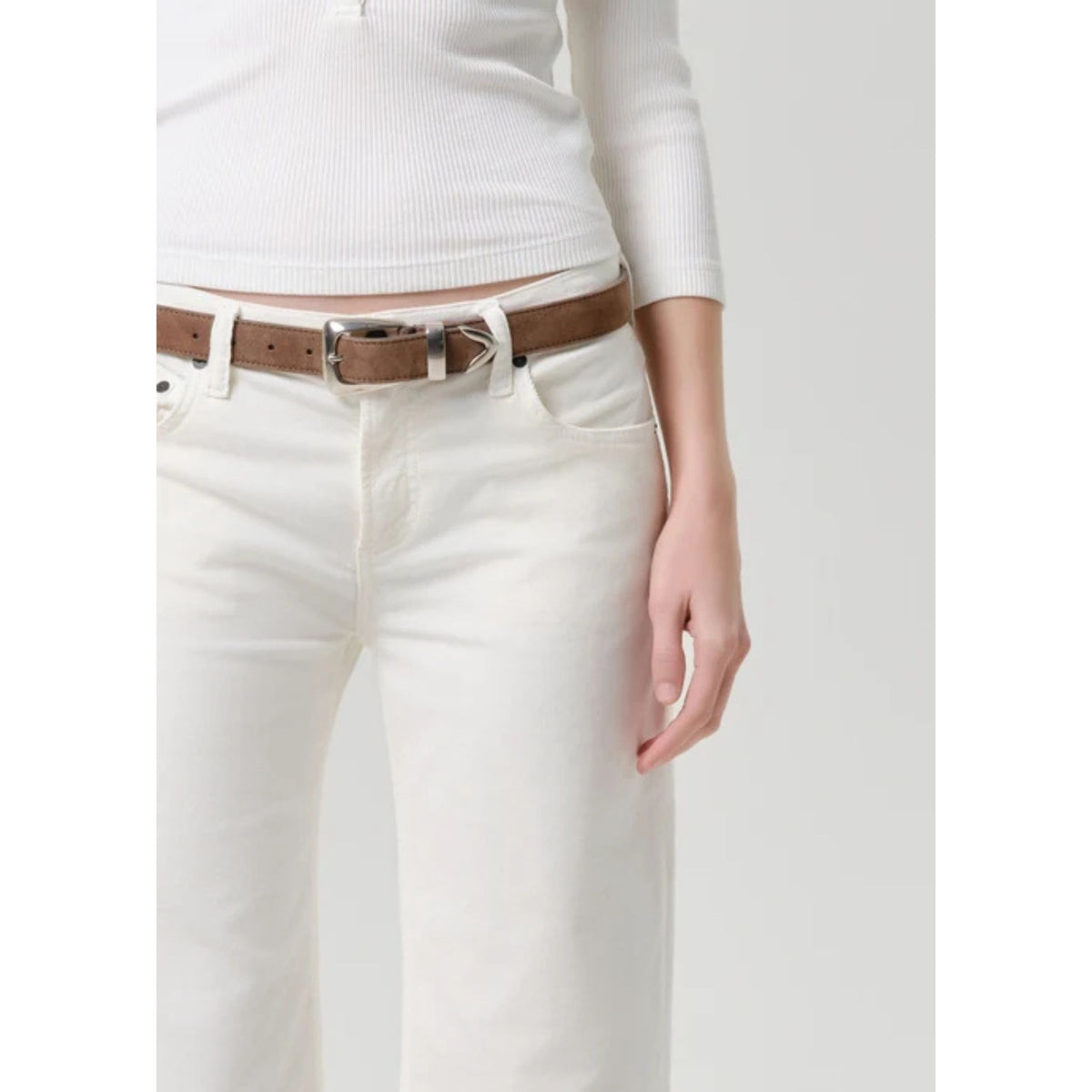 A close-up of a person wearing white jeans and a brown leather belt with a silver buckle, paired with a white ribbed top against a light gray background.