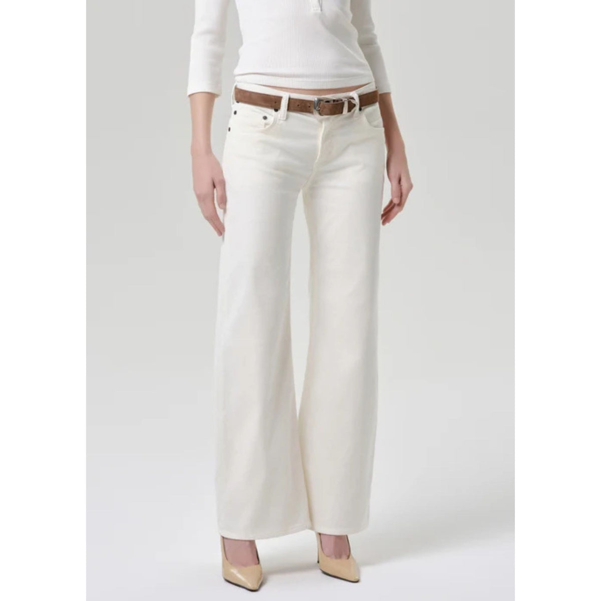 A model wearing off-white wide-leg jeans with a brown belt, paired with a white long-sleeve top and beige pointed-toe heels, shown against a light gray studio background.