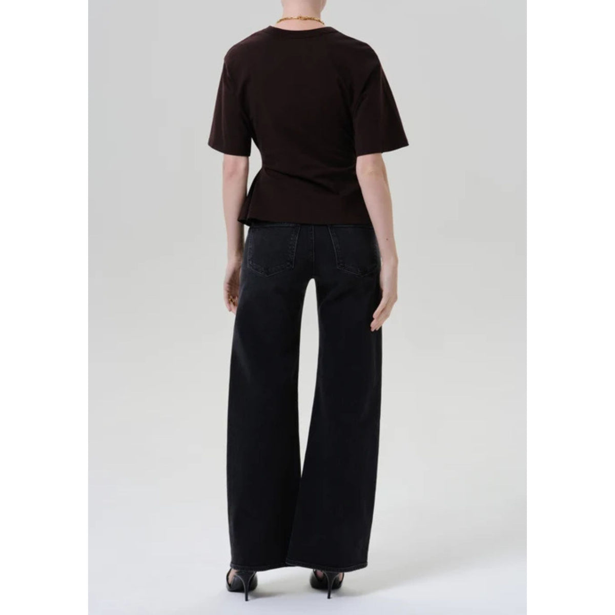 A model shown from the back wearing a dark brown short-sleeve t-shirt with a peplum waist and black wide-leg jeans, set against a plain light gray studio background.