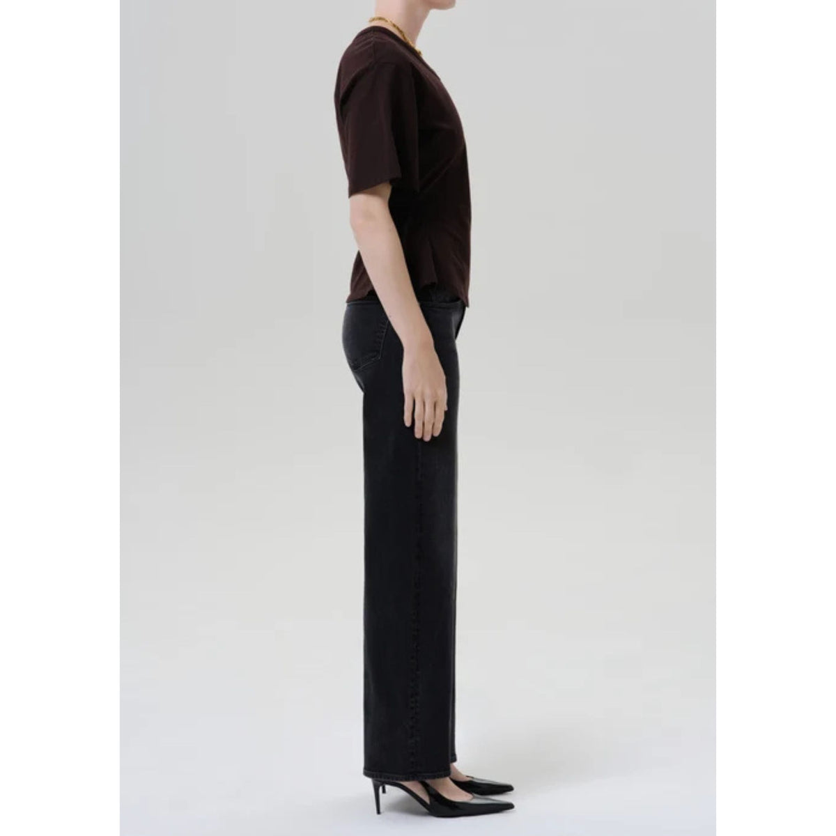 A side-profile studio shot of a model wearing a dark brown short-sleeve top, black wide-leg pants, and black pointed-toe heels against a light gray background.