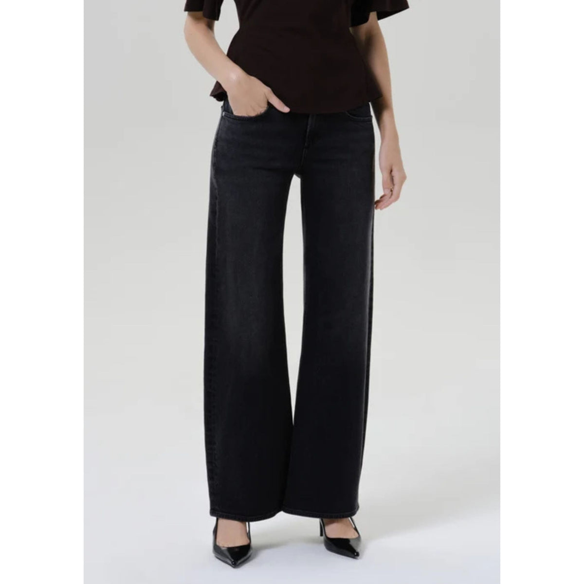 A model wearing black wide-leg denim jeans, a dark brown ruffled top, and black pointed-toe heels, standing against a light gray studio background.