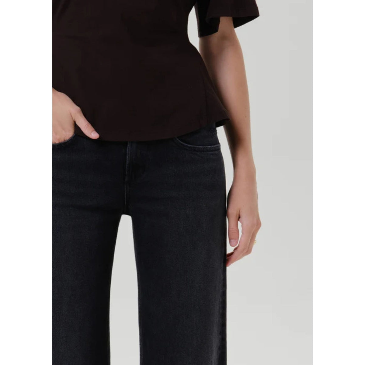 A close-up of a person wearing dark charcoal gray slim-fit jeans and a dark brown short-sleeve top, cropped from the chest down against a light gray studio background.