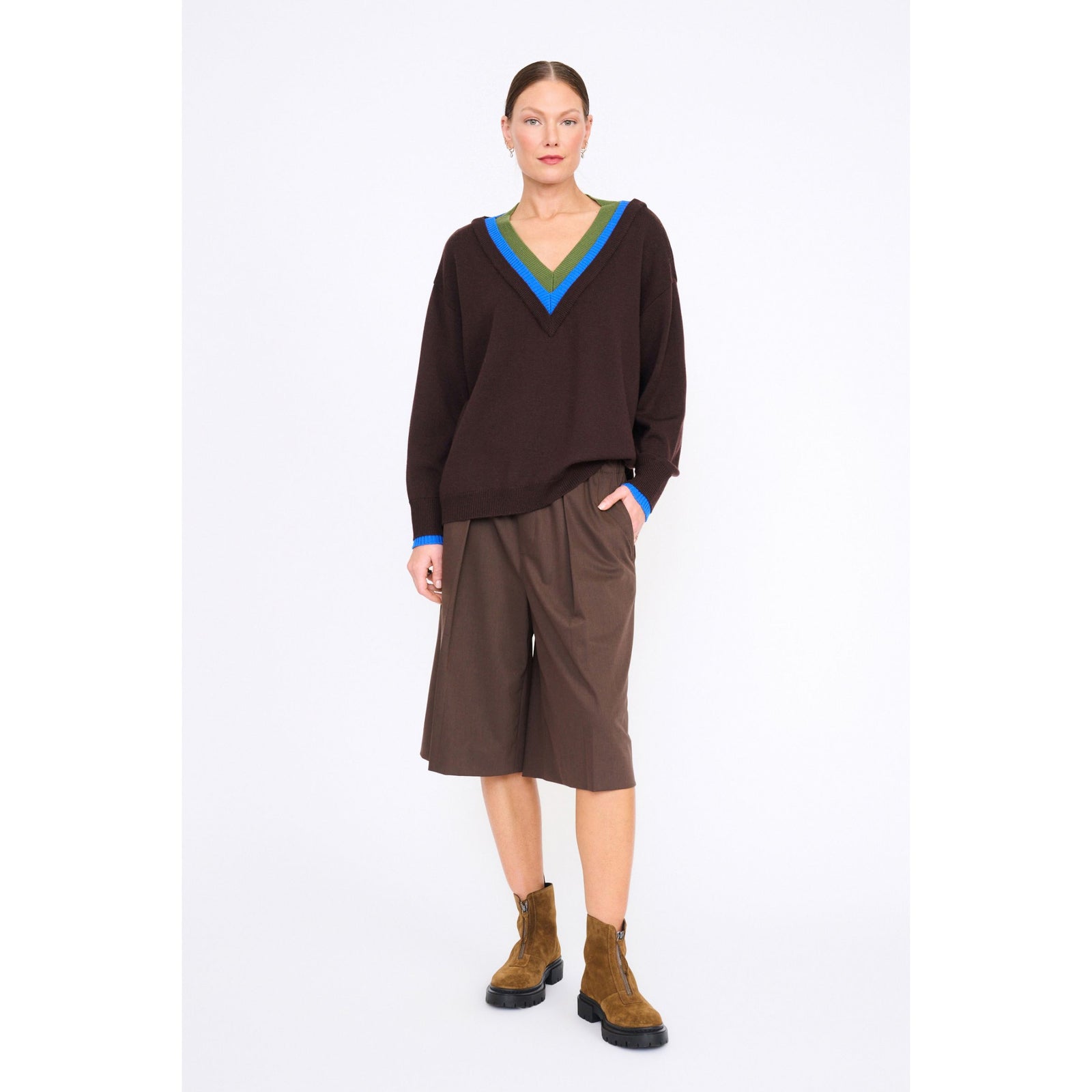 A model stands against a plain white studio background wearing a dark brown long-sleeve V-neck sweater with bright blue and olive green trim, matching brown shorts, and brown suede ankle boots.
