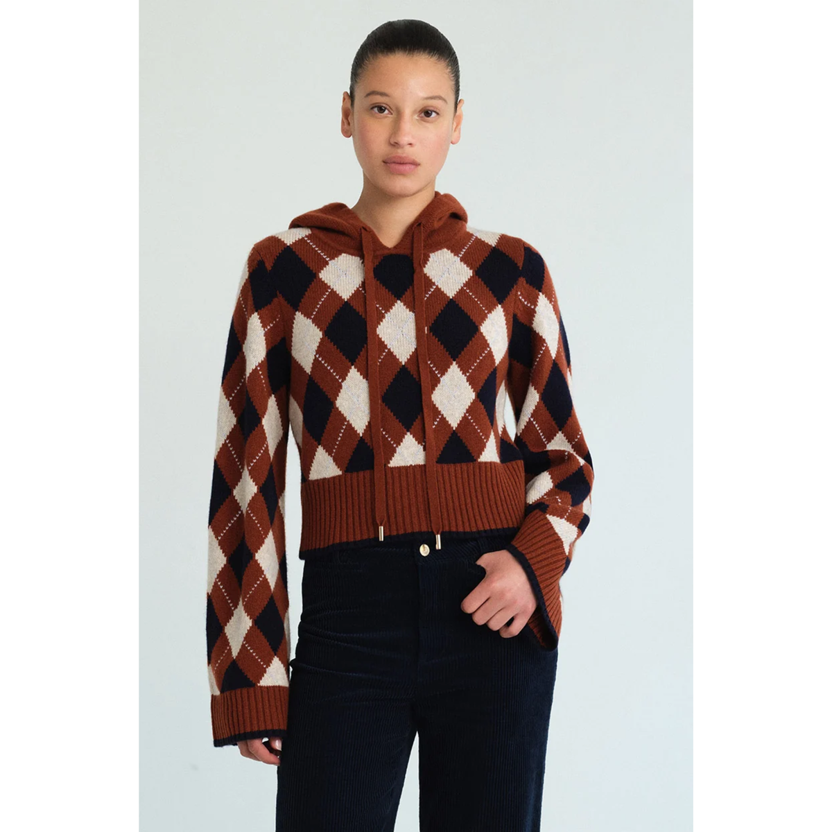 A model wearing a cropped, long-sleeve argyle knit hoodie in rust brown, cream, and black, paired with dark pants, against a light gray studio background.