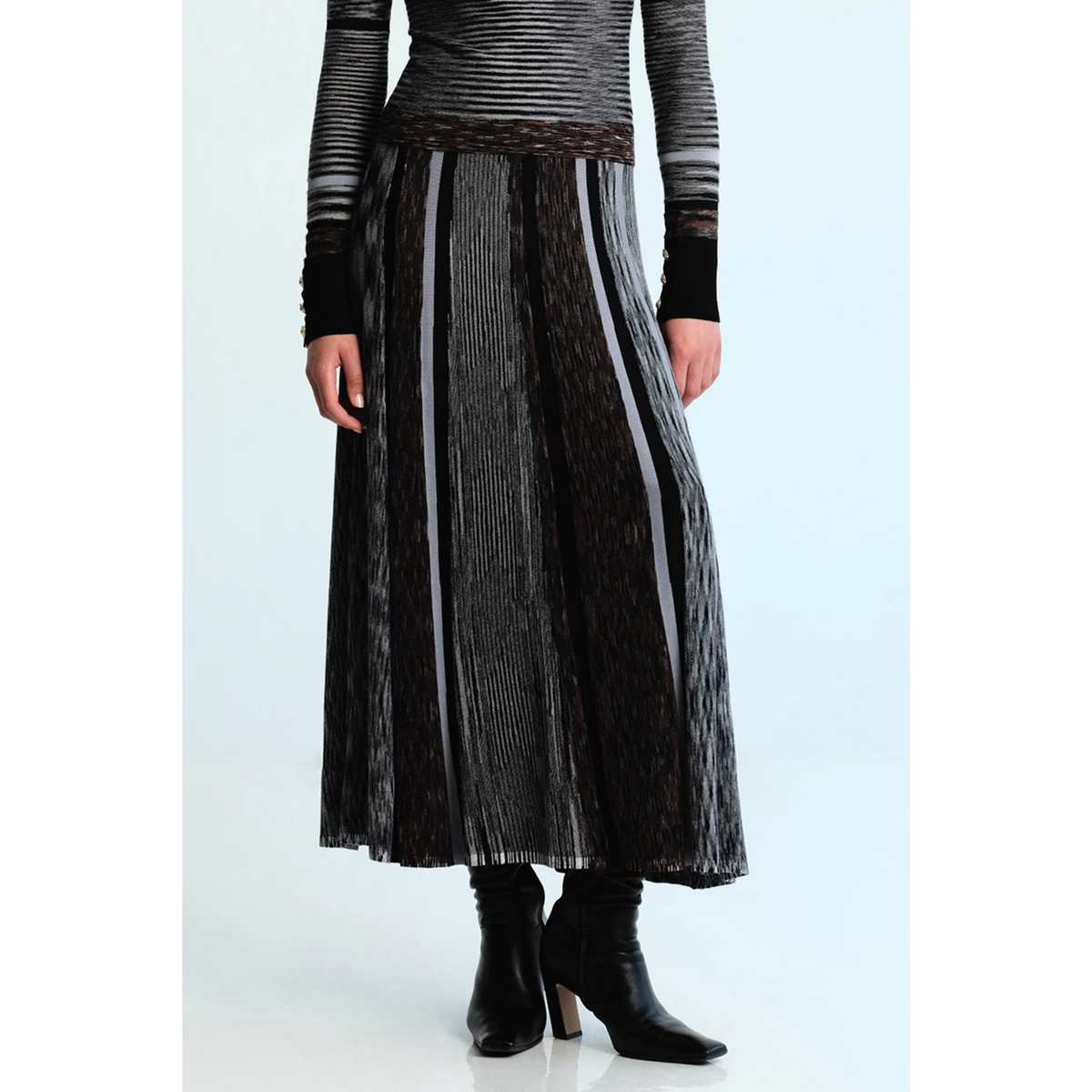 A model wearing a long, pleated midi skirt in black and gray with vertical stripes, paired with a matching striped top and black ankle boots, set against a light blue studio background.