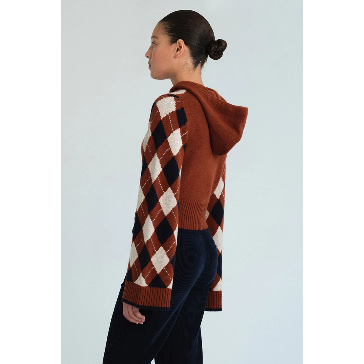 A woman shown from the side profile wearing a brown, cream, and black argyle patterned hooded sweater, paired with dark navy pants, against a light gray studio background.