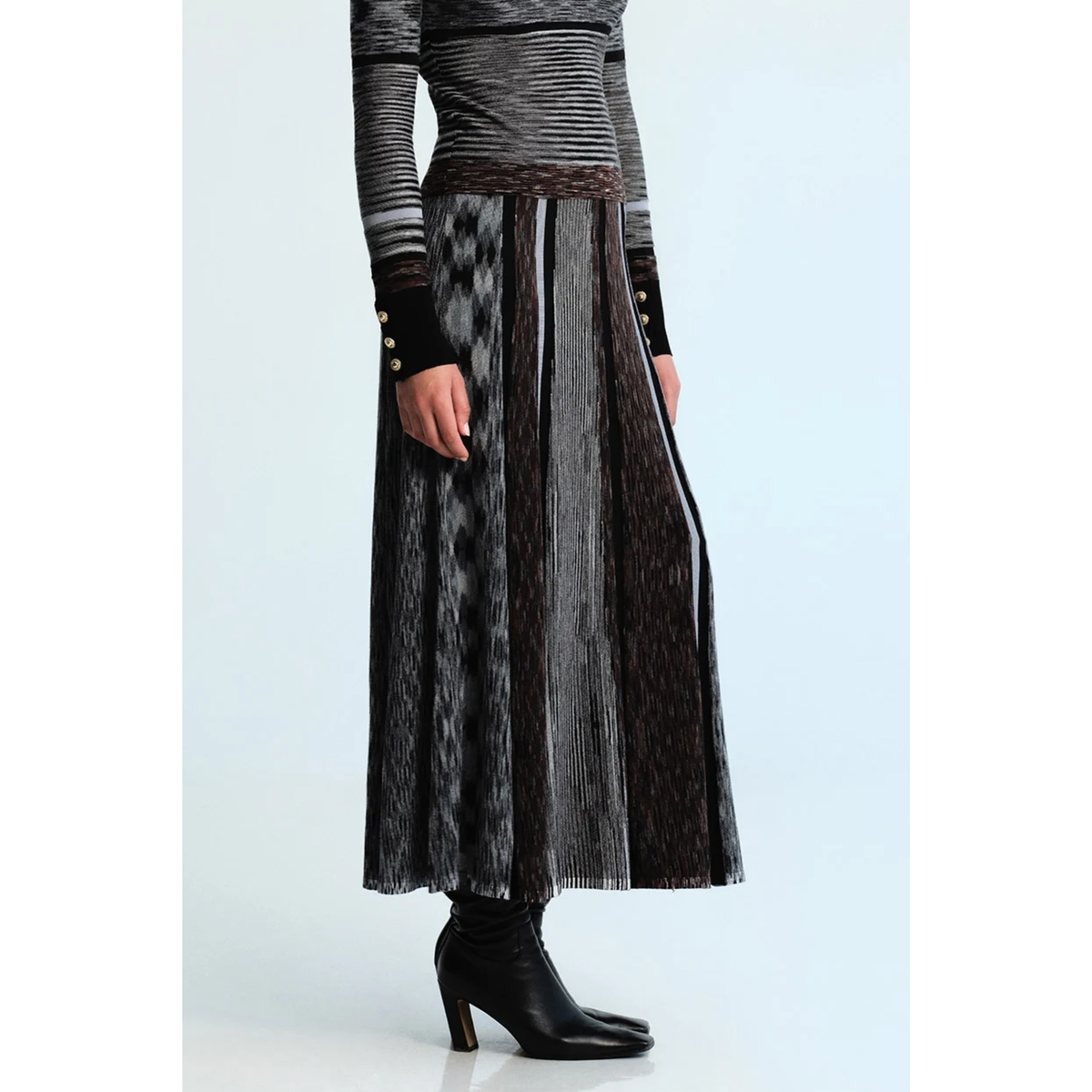 A model wearing a long, pleated midi skirt with vertical textured stripes in black, gray, and brown, paired with a striped top and black ankle boots, set against a light blue studio background.