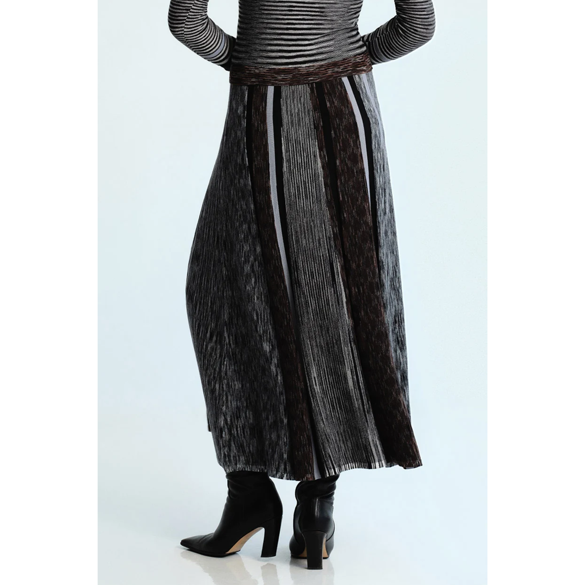 A model is shown from the back wearing a long, pleated skirt with vertical stripes in black, gray, and brown, paired with a matching striped top and black ankle boots.