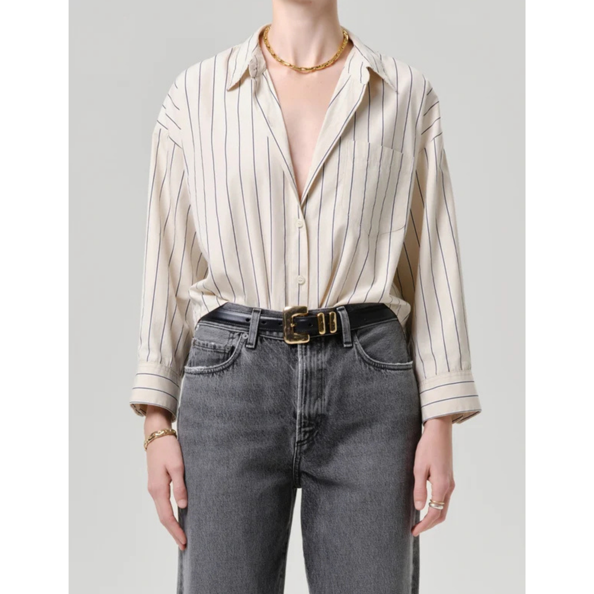 A model wearing a cream-colored long-sleeve button-up shirt with thin vertical black stripes, tucked into dark gray jeans with a black belt, against a solid light gray background.