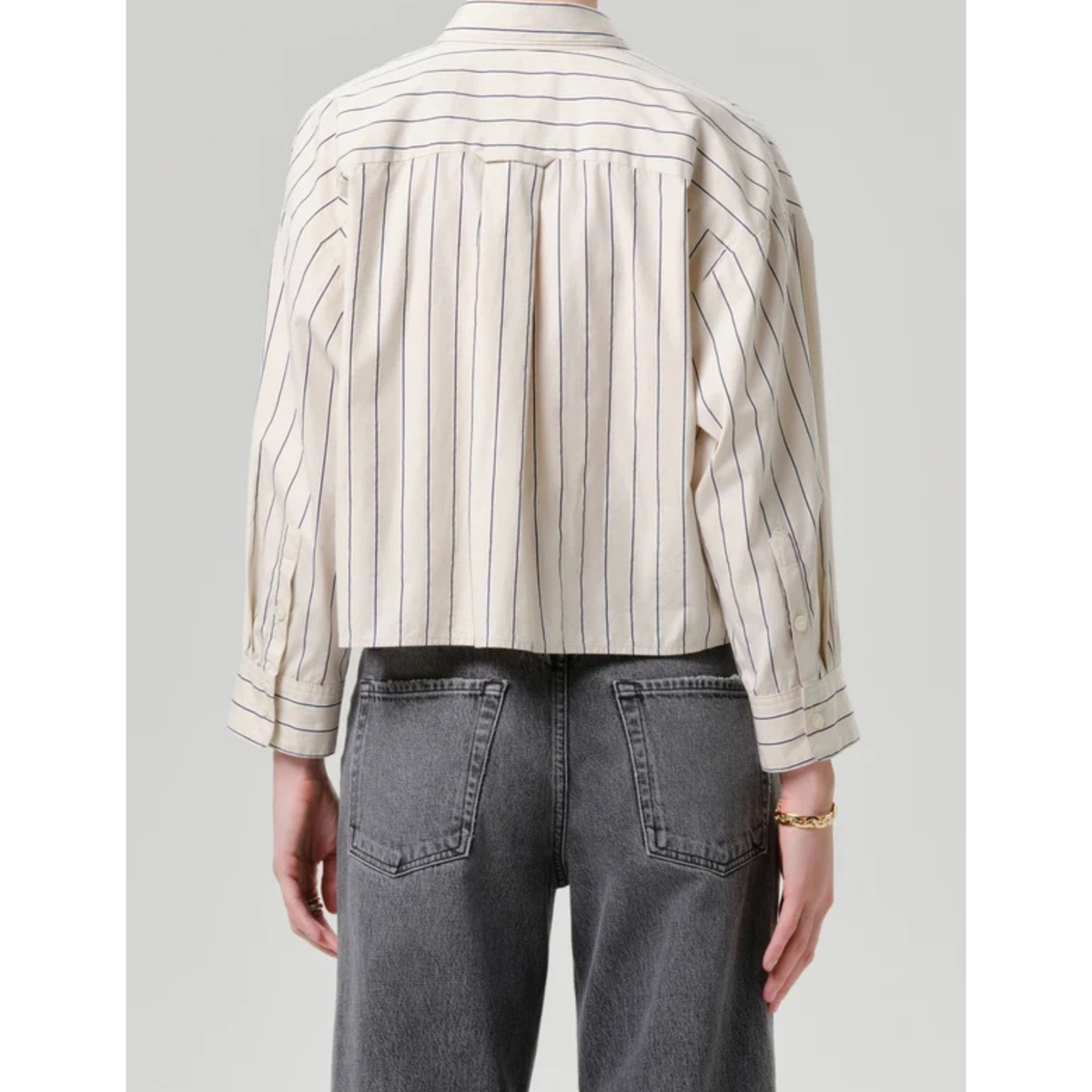 Back view of a person wearing a cropped, long-sleeve striped shirt with vertical thin stripes on an off-white base, paired with dark gray jeans, against a light gray studio background.