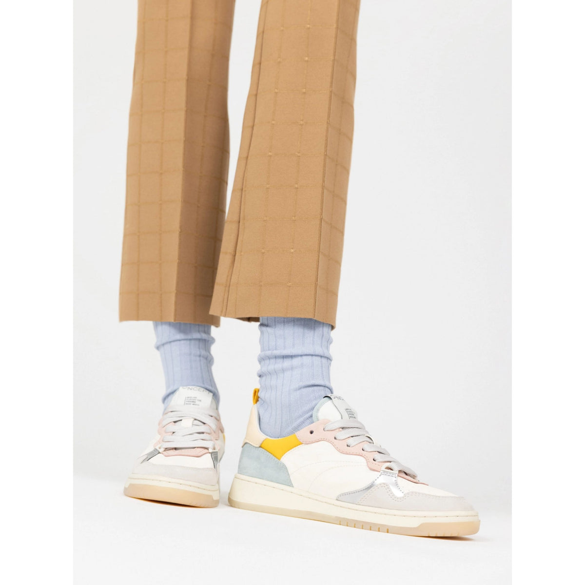 A person wearing tan plaid cropped trousers, light blue ribbed socks, and multi-colored low-top sneakers in cream, pink, light blue, and yellow, standing against a plain white studio background.