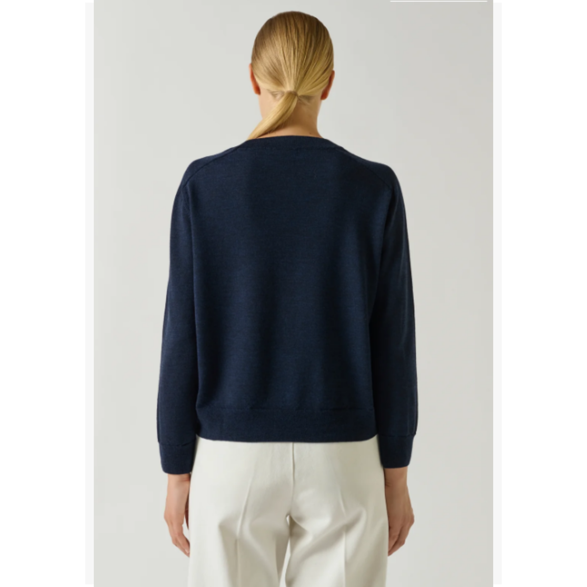 A woman viewed from the back wearing a dark navy long-sleeve knit sweater and off-white trousers, set against a plain light gray studio background.