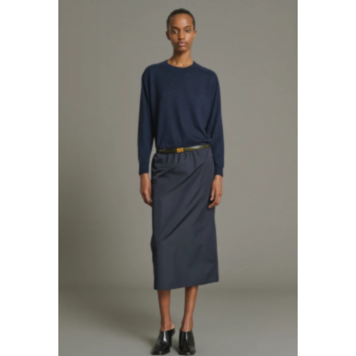 A model stands against a plain gray studio background wearing a navy blue long-sleeve top and a matching navy midi skirt with a thin black belt featuring a gold buckle.