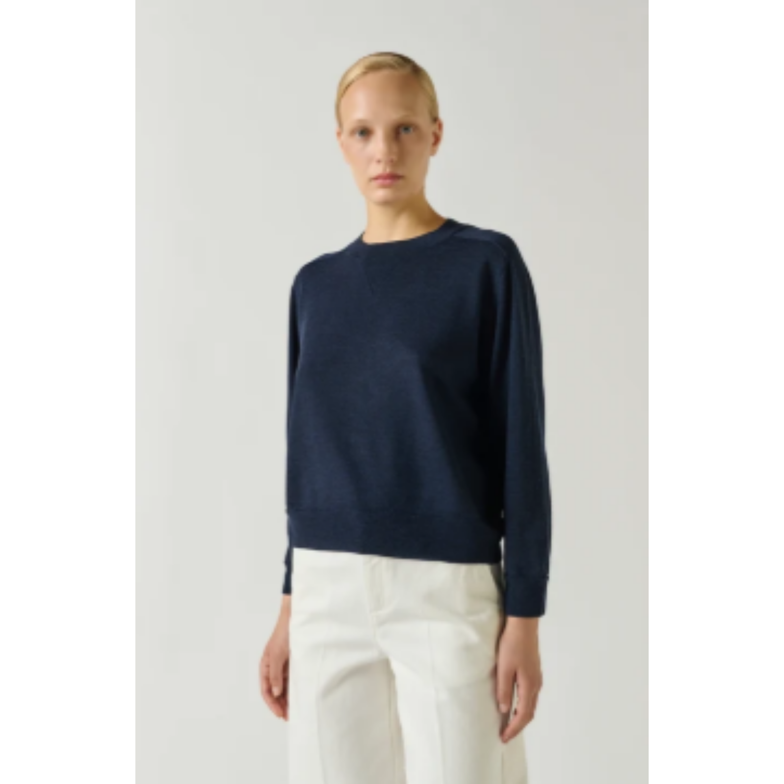 A woman modeling a navy blue long-sleeve knit sweater with white trousers, standing against a light gray studio background.