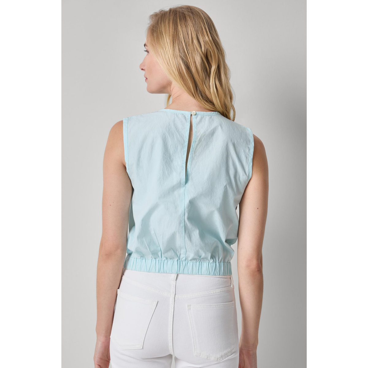 Woman wearing a light blue sleeveless top and white pants on a gray background