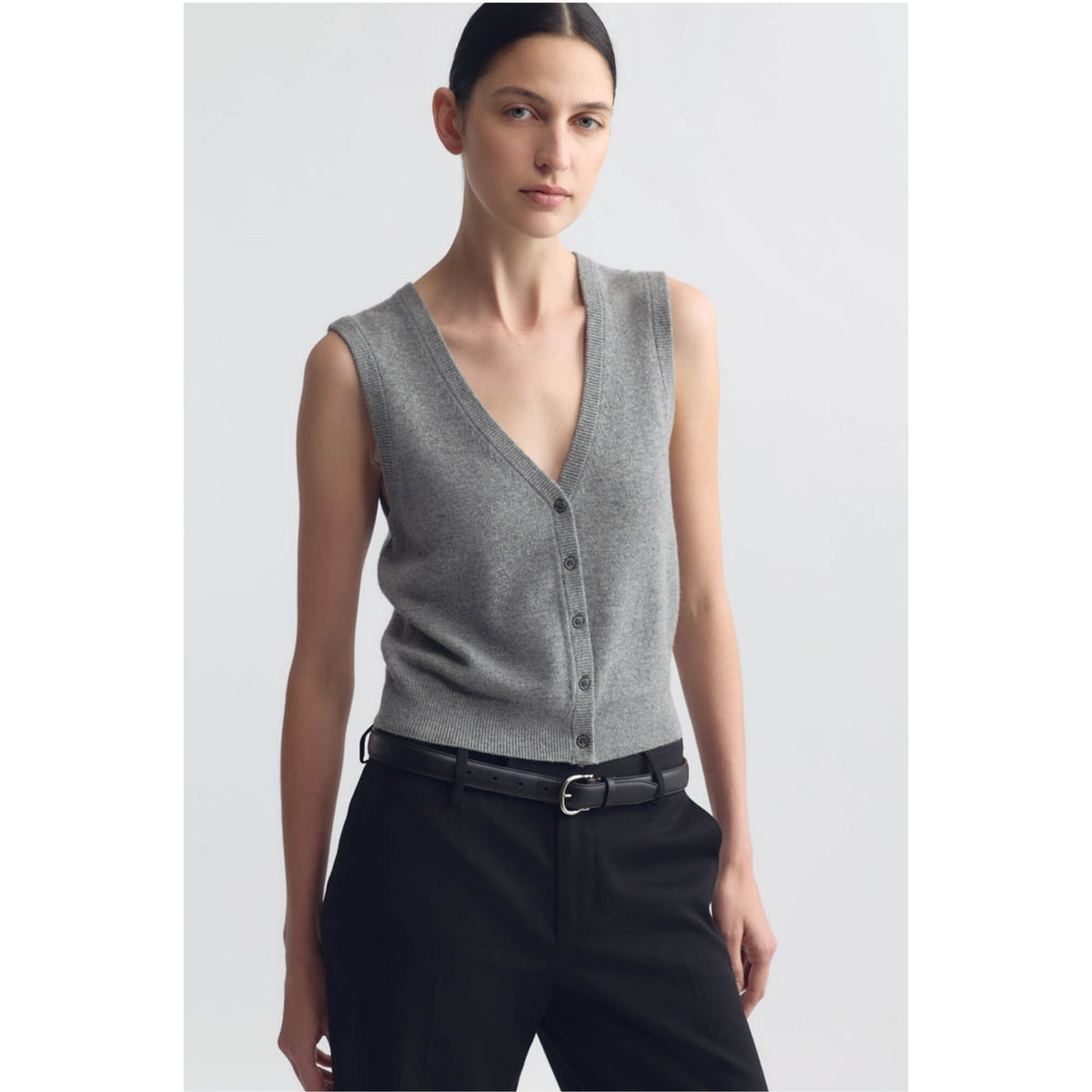 A model wearing a gray sleeveless V-neck knit vest with front buttons, paired with black trousers and a black belt, against a light gray studio background.
