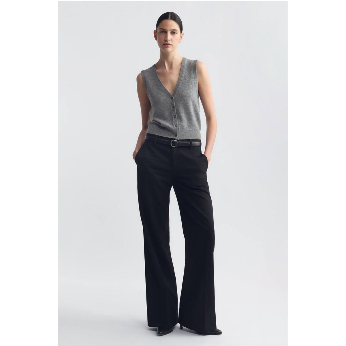 A model stands against a light gray studio background wearing a sleeveless gray knit vest with a V-neck and front buttons, paired with black wide-leg trousers and a black belt.
