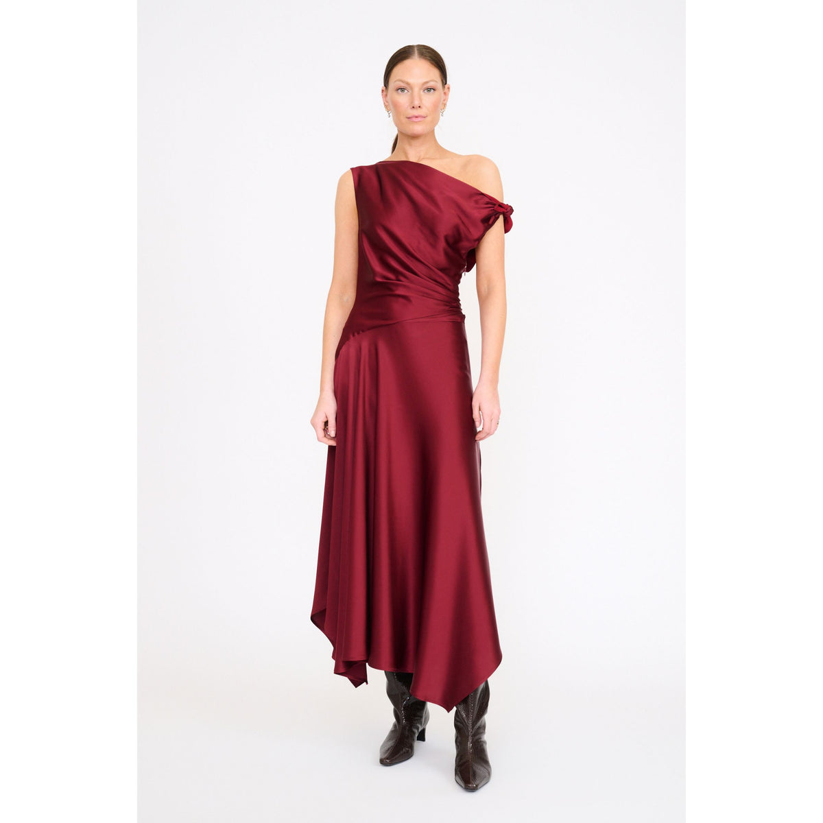 A woman modeling a satin-like one-shoulder midi dress in deep burgundy, paired with dark brown boots, against a plain white studio background.