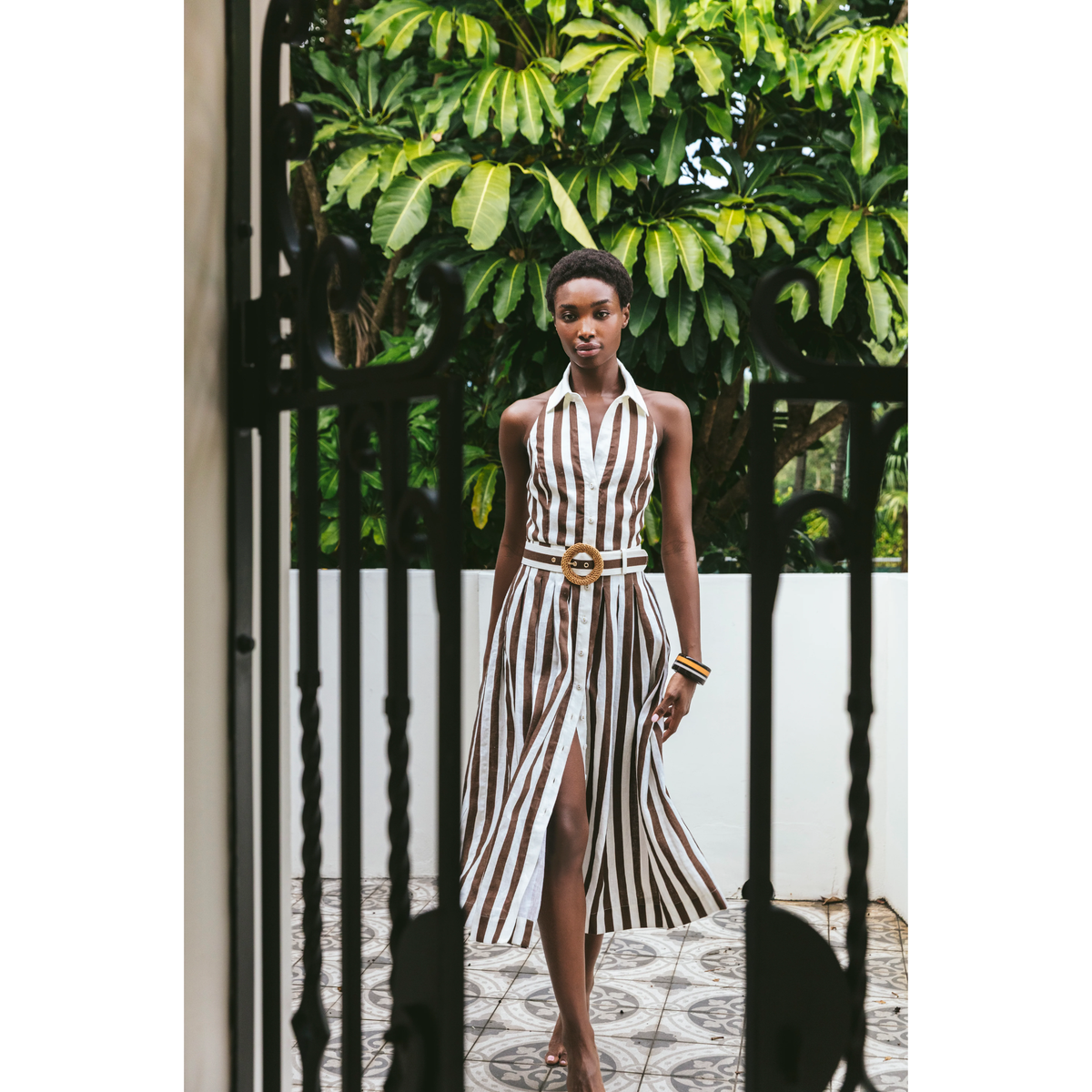 A woman models a sleeveless halter-neck midi dress with vertical brown and white stripes, featuring a matching belt with a gold buckle, set against a background of green foliage and a black metal gate.