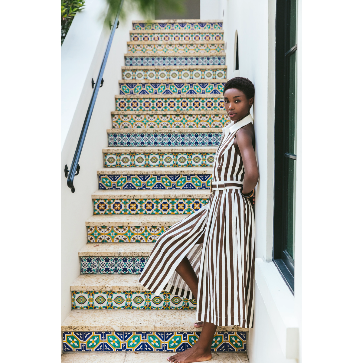 A woman wearing a sleeveless brown and cream striped midi dress with a matching belt stands on a set of tiled outdoor stairs.
