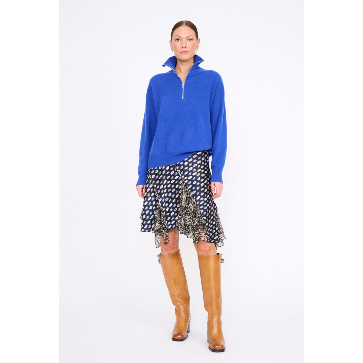 A model stands against a light gray studio background wearing a bright blue long-sleeve quarter-zip pullover, a black and white patterned ruffled skirt, and tall tan leather knee-high boots.